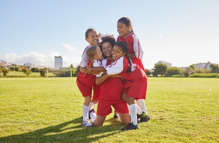 A girls soccer team stands on the field sharing a group hug